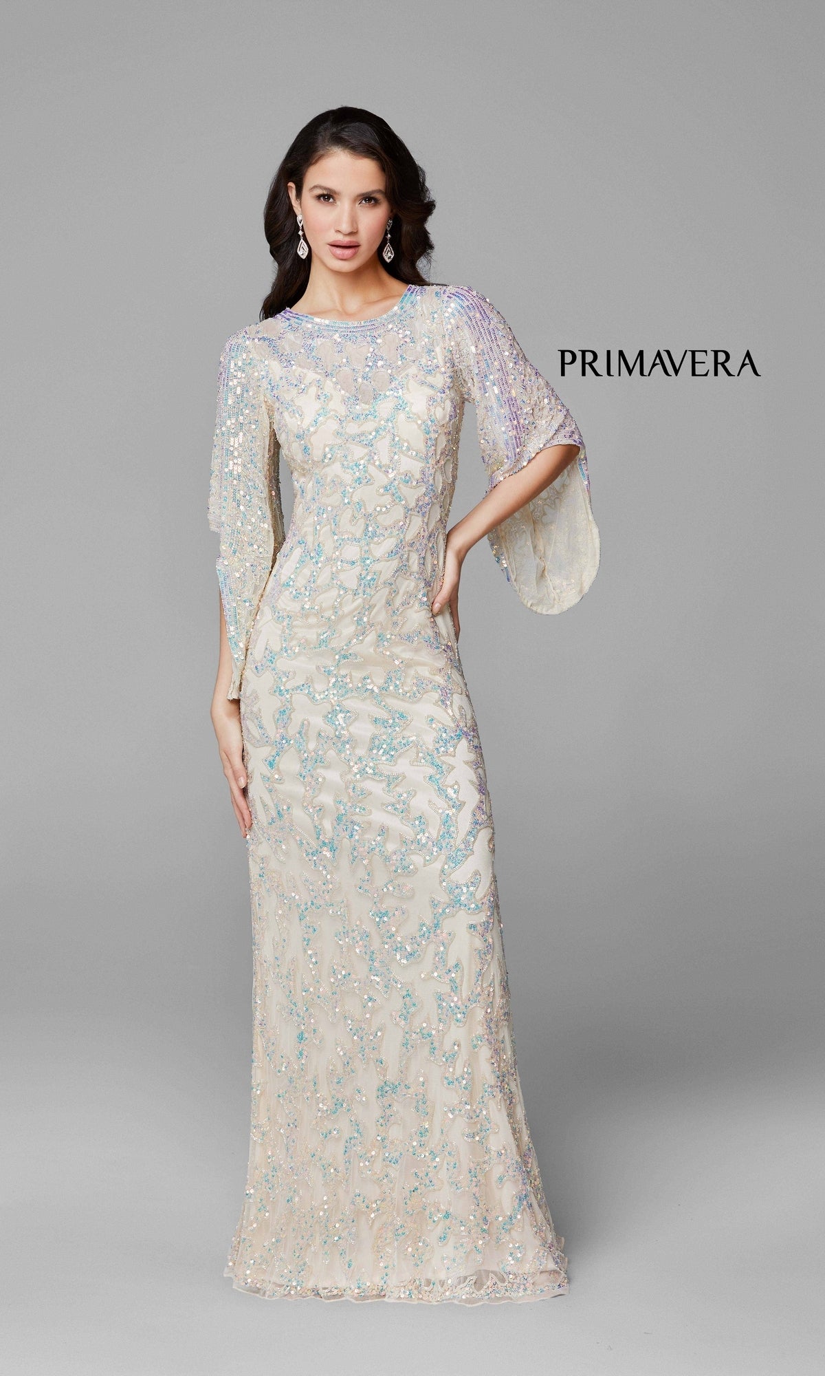 A woman in the Long Bell Sleeve Beaded Evening Gown 9713, featuring ivory fabric, light blue sequins, and sheer flutter sleeves, stands against a gray background with